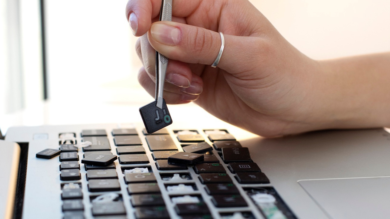 Una mano di una donna che tiene con una pinzetta un tasto sopra una tastiera di laptop da cui mancano numerosi tasti.