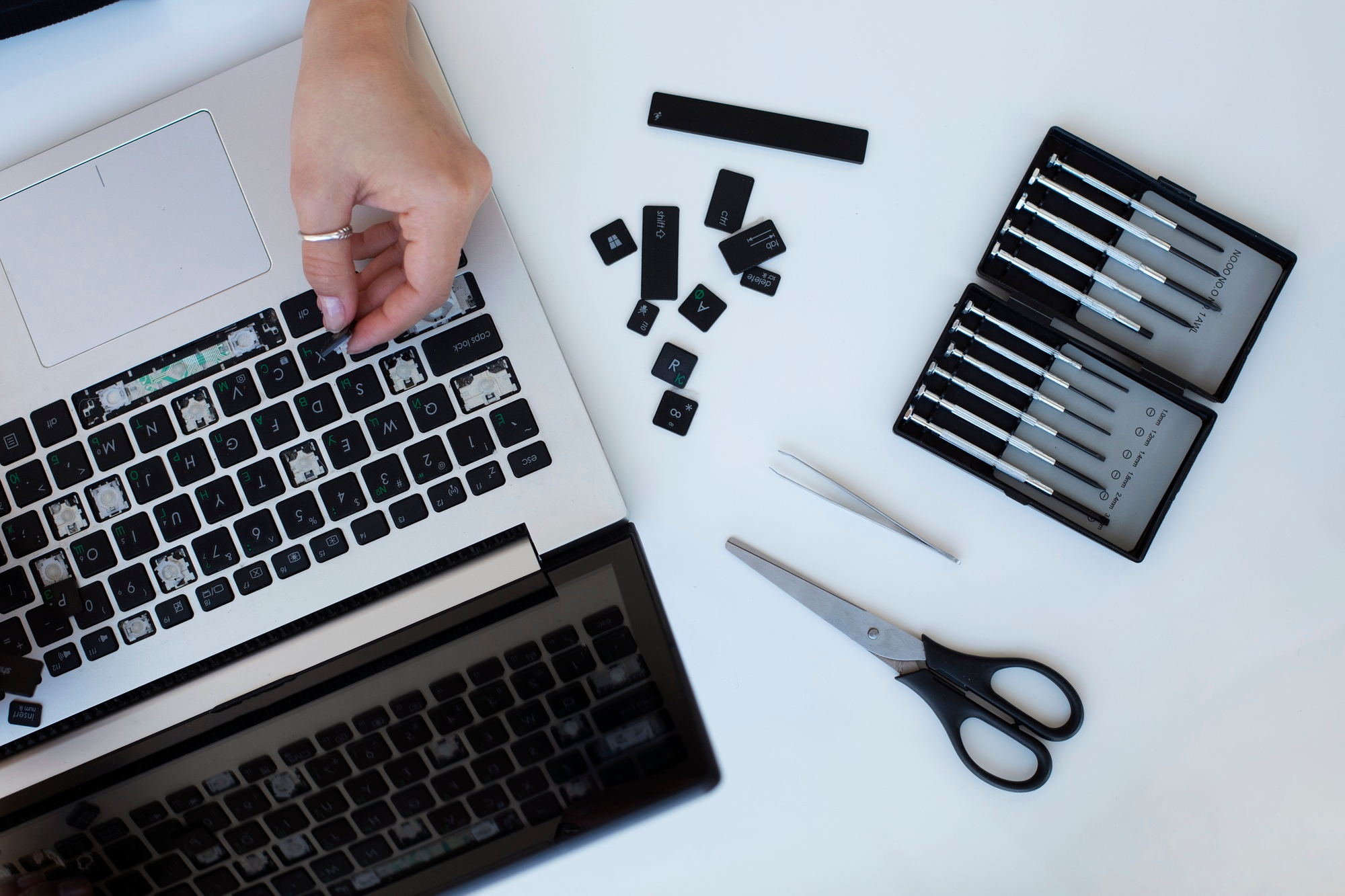 Una tastiera di laptop con tasti rimossi, strumenti e la mano di una persona che lavora alla sua riparazione.